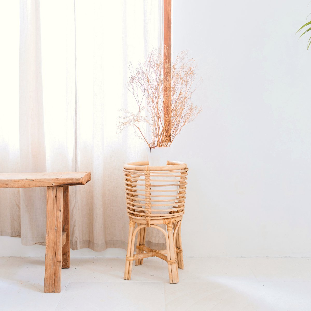 Rotan plantenstandaard met een witte bloempot met droogbloemen erin naast een houten bankje