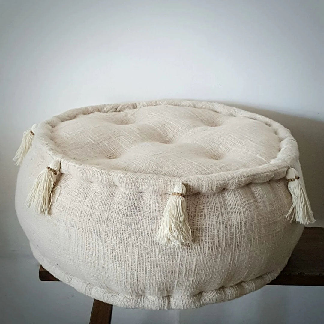 Kleine ronde poef in de kleur beige op een houten bankje tegen een muur aan, zijkant en voorkant zichtbaar