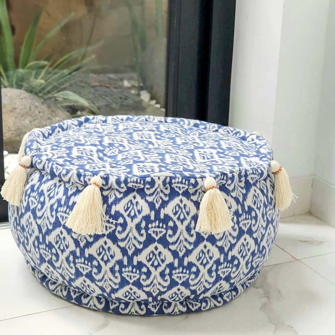 Kleine ronde blauw witte poef van katoen met witte kwastjes aan de zijkant op de grond voor een raam