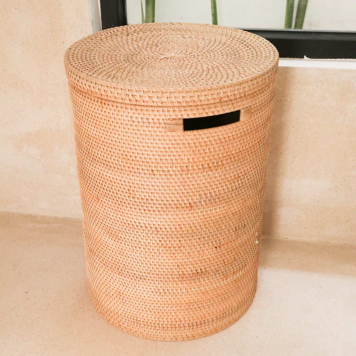 Rotan wasmand met deksel en handvaten staand voor een raam