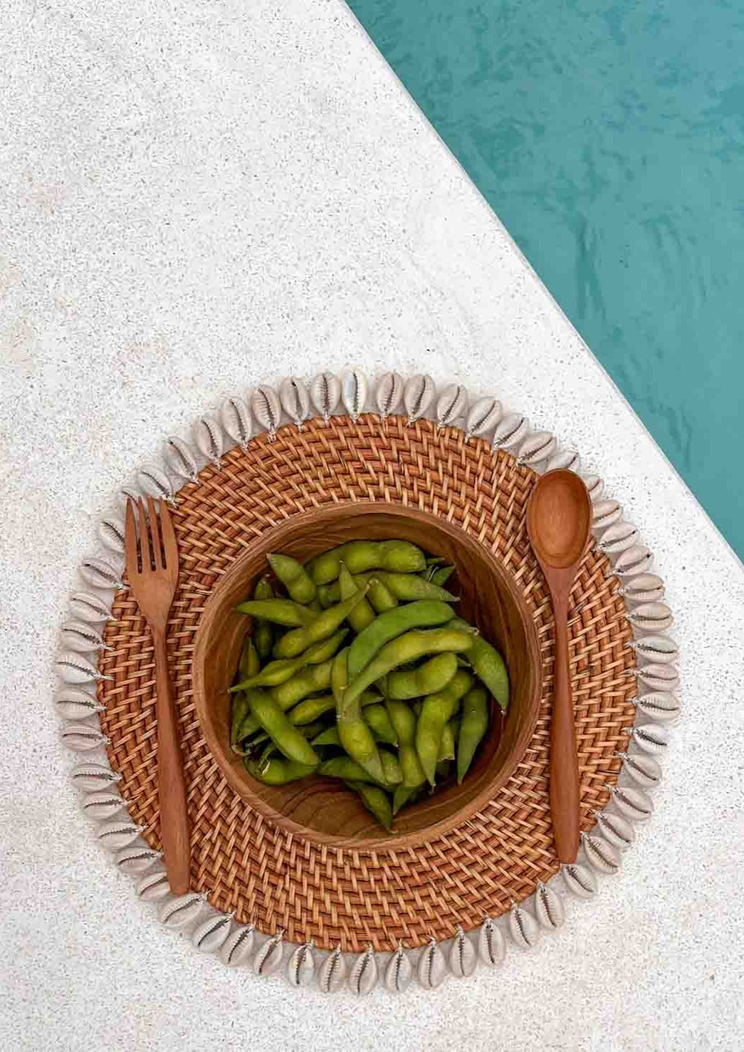 Schelpen placemat met daarop houten bestek en een houten kom naast een zwembad