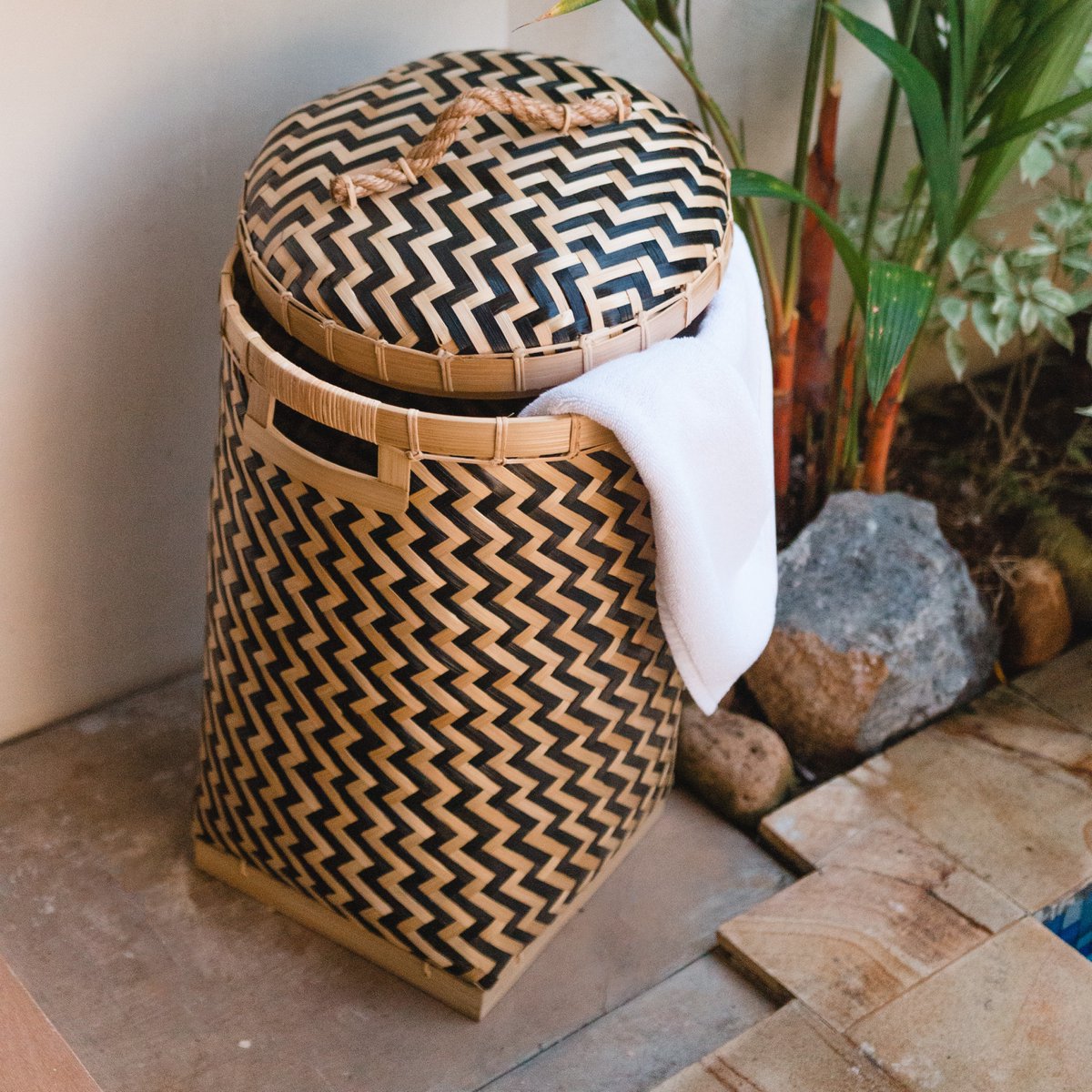 Wasmand van bamboe met een deksel erop en een handdoek erin naast een plant