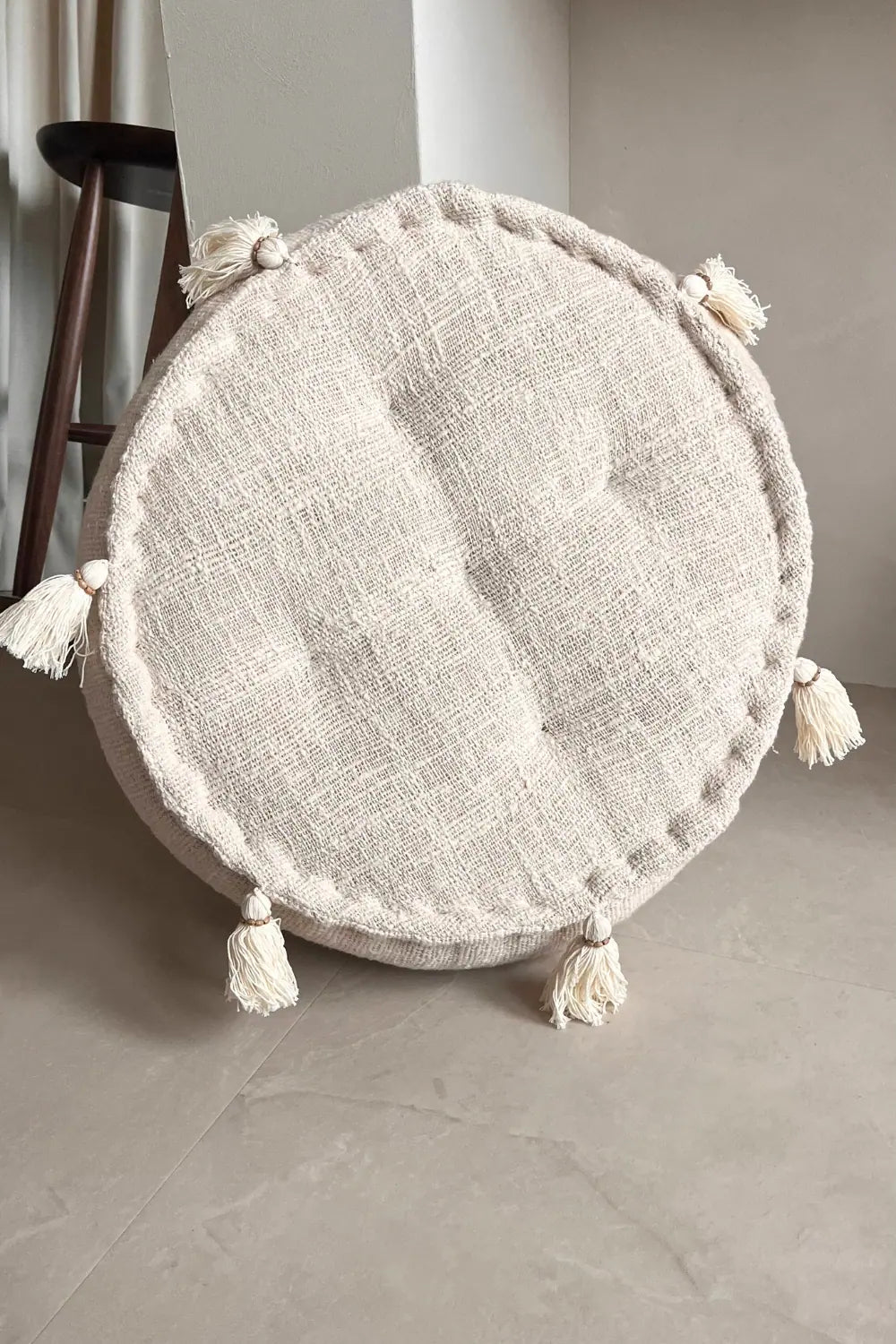 Ronde poef van katoen in creme kleur met franjes rechtop staand tegen een muur 