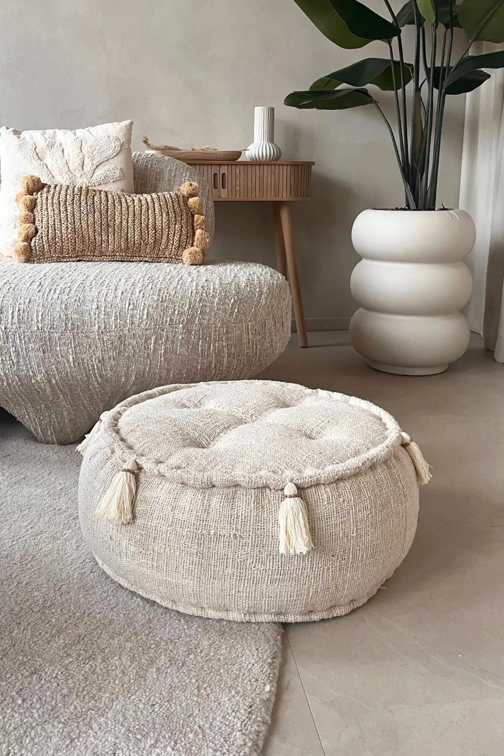 Ronde poef van natuurlijk katoen in creme kleur staand op een vloerkleed voor een bank