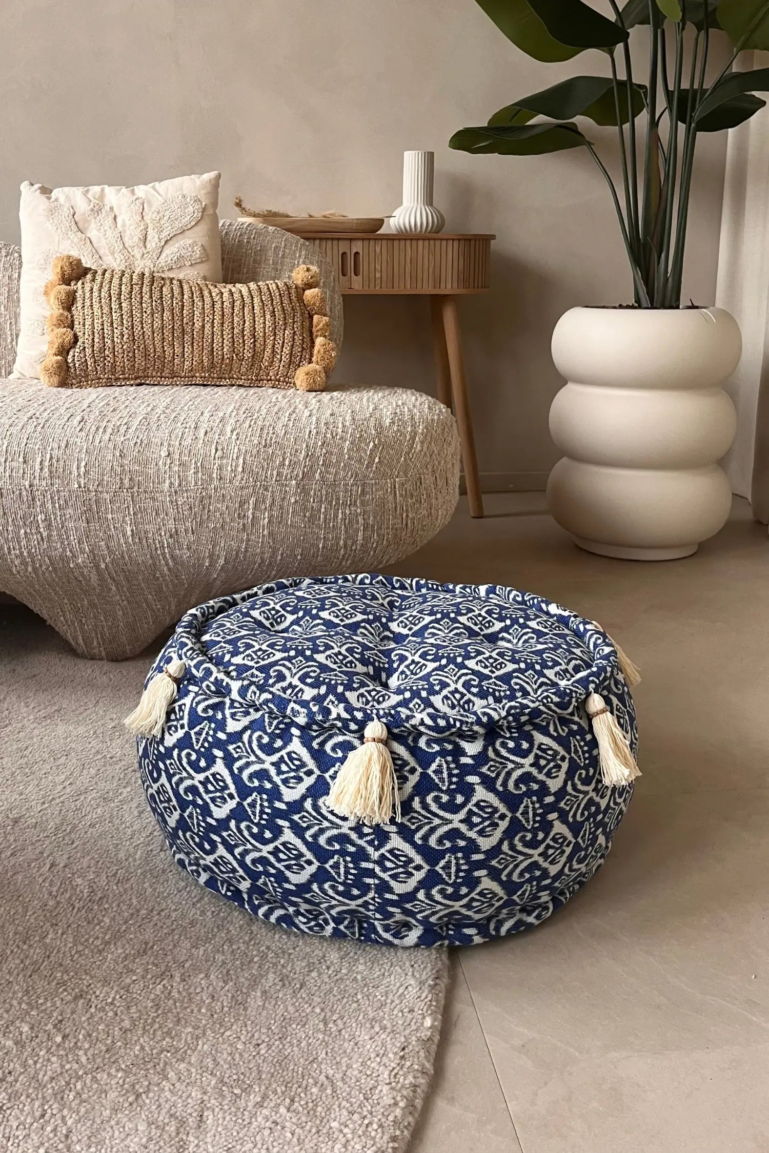 Ronde blauw witte poef met franjes liggend op een vloerkleed voor een bank