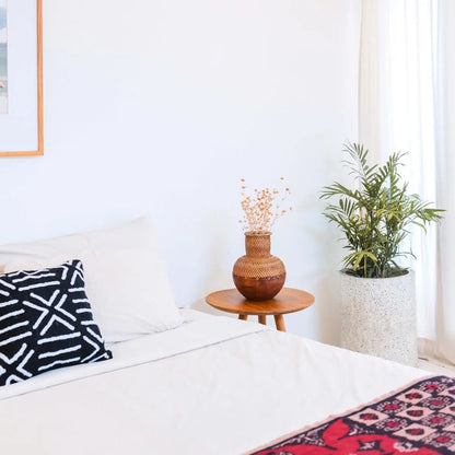 Vaas van rotan en hout met bloemen erin op een tafeltje naast een bed 