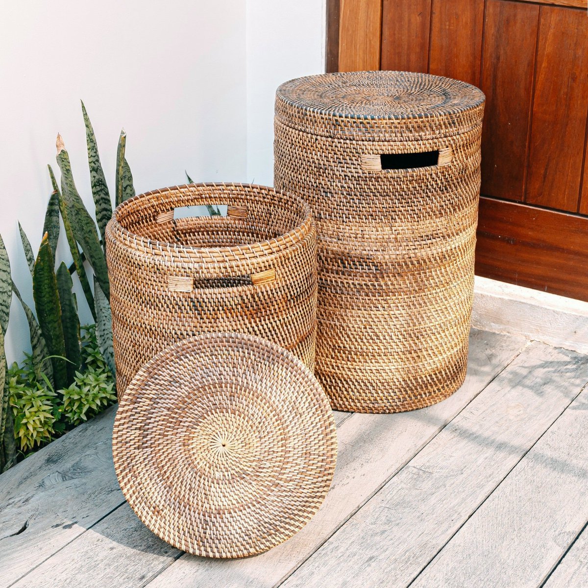 Twee ronde rotan wasmanden met deksel in bruine tinten, buiten naast planten geplaatst.