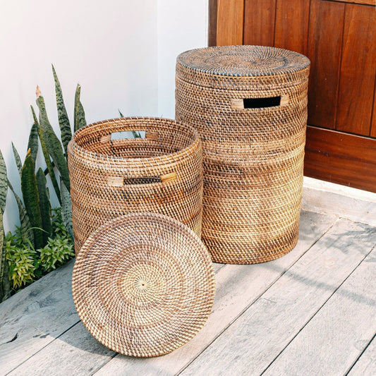 Twee ronde rotan wasmanden met deksel in bruine tinten, buiten naast planten geplaatst.