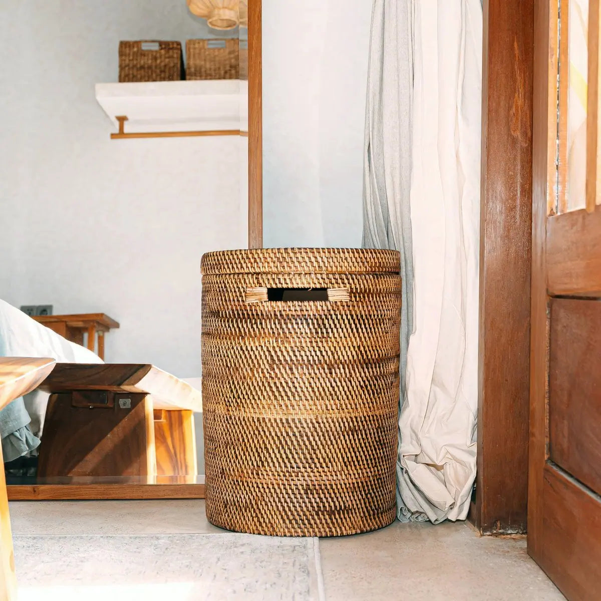 Rotan wasmand met deksel in een lichte kamer bij een houten deur, staand naast een gordijn