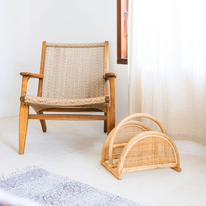 Rotan tijdschriftenmand op de vloer naast een houten stoel met rotan zitting en rugleuning, in een lichte kamer.